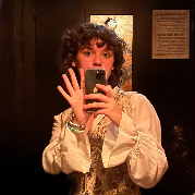 a young white person with brown hair brown eyes.they are standing in a dimly lit resteraunt bathroom taking a mirror photo. they are only visible from the waist up. their hair is in a shag with bangs and their mouth is covered by their phone. he is wearing a flowy long-sleeve shirt and a floral patterend vest.